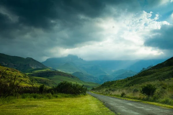 空の息をのむような雲の下で草で覆われたフィールドや丘に囲まれた道路 — ストック写真