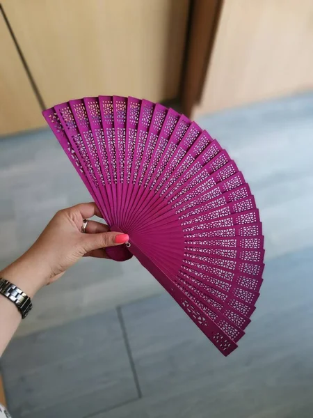 A purple folding hand fan held by woman\'s hand with pink nails and a watch