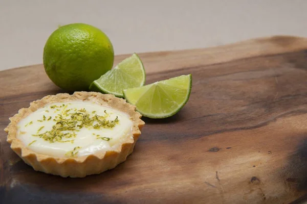 A closeup of a key lime tartlet on a wooden board with key lime fruits