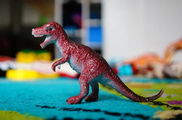 A closeup of a red roaring toy dinosaur on a blue carpet under the lights with a blurry background