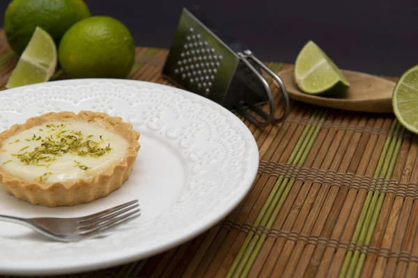 A soft focus of a key lime tartlet and a fork on a plate with key lime fruits and grater in the background on a bamboo mat