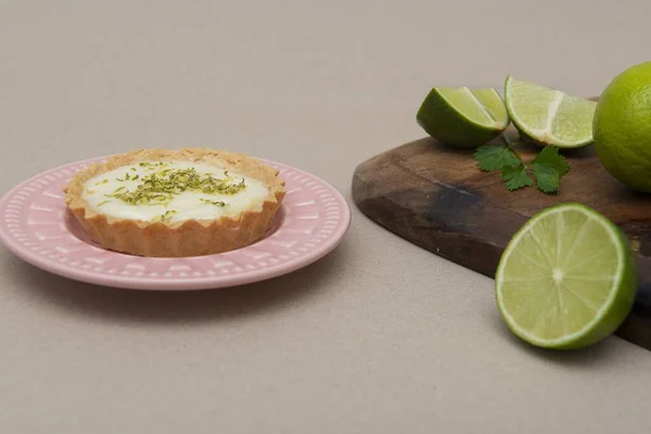 A closeup of a key lime tartlet on a pink plate with some sliced key limes on a wooden board
