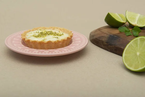 A closeup of a key lime tartlet on a pink plate with key lime fruits on a wooden board