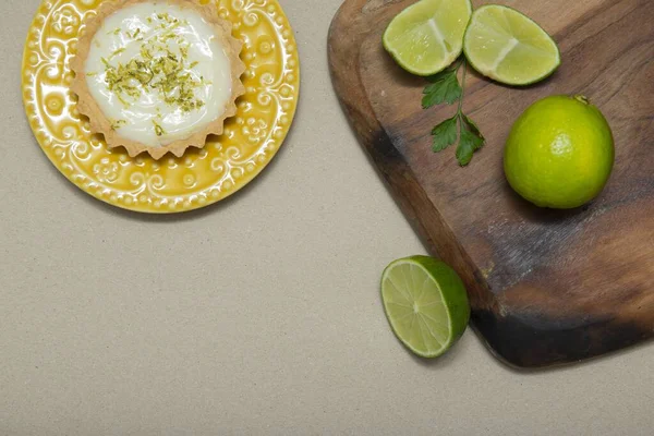 A top view of a key lime tartlet on a plate with key lime fruits on a wooden board