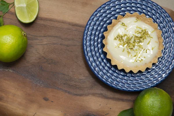 A top view of a key lime tartlet on a blue plate and key lime fruits on a wooden board