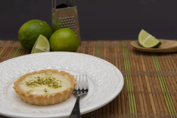 A soft focus of a key lime tartlet and a fork on a plate with key lime fruits and grater in the background on a bamboo mat