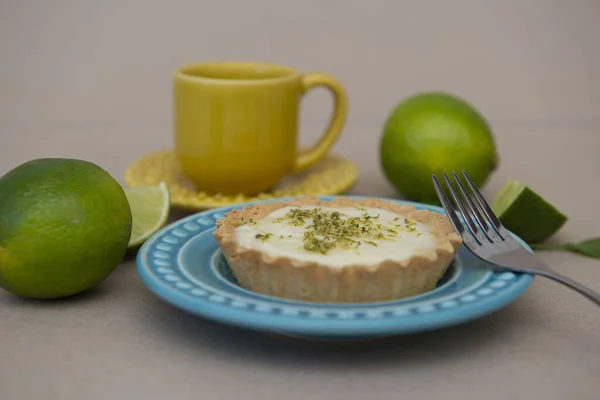 A soft focus of a key lime tartlet and a fork on a plate and key lime fruits and a yellow mug in the background