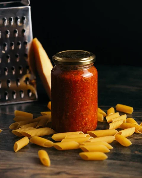 A vertical shot of penne pasta and a jar of sauce on the table with cheese and a grater on the background