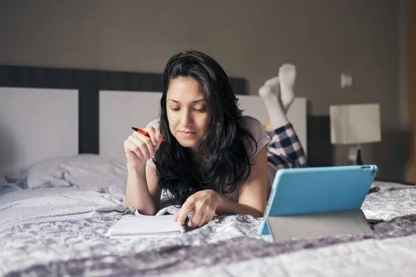 여성이 노트북으로 집에서 침대에 누워서 타블렛으로 인터넷을 공부하고 있습니다 — 스톡 사진