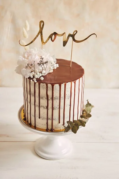 白い背景の白いテーブルの上にチョコレートドリップと愛のトッパーとおいしいウェディングケーキの垂直クローズアップショット — ストック写真
