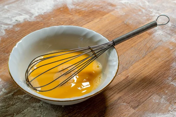小麦粉で覆われたテーブルの上に置く泡立て器で溶いた卵の白いボウル — ストック写真