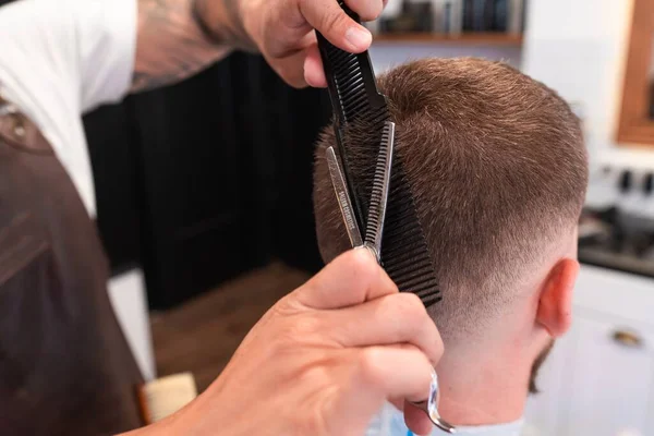 理发店里一个年轻理发师用梳子和剪刀剪短男顾客头发的特写镜头 — 图库照片
