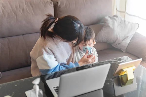 若いアジアの母親を置く彼女の子供の顔マスク上のCovid 19予防のために自宅で作業しながら — ストック写真