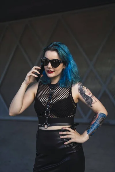 建物の側面に電話を介して話してサングラスとセクシーな黒のドレスを身に着けている入れ墨と青い髪を持つユーロ圏の女性 — ストック写真