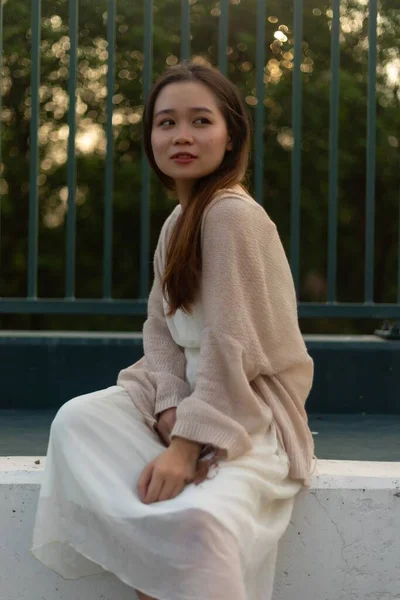 金属製のフェンスの近くに座ってジャケットと白いドレスを着て長い髪を持つ魅力的なアジアの若い女性 — ストック写真