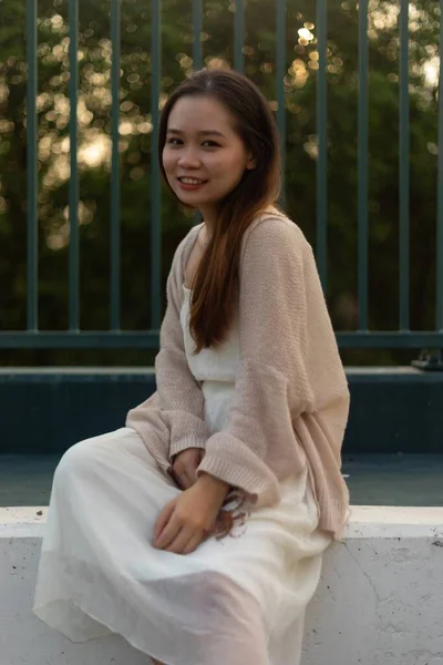 金属製のフェンスの近くに座ってジャケットと白いドレスを着て長い髪を持つ魅力的なアジアの若い女性 — ストック写真