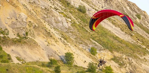 A tandem of people under the red dome of a motor paraglider takes off into the air in a mountain gorge. Red paraglider on the background of lime mount