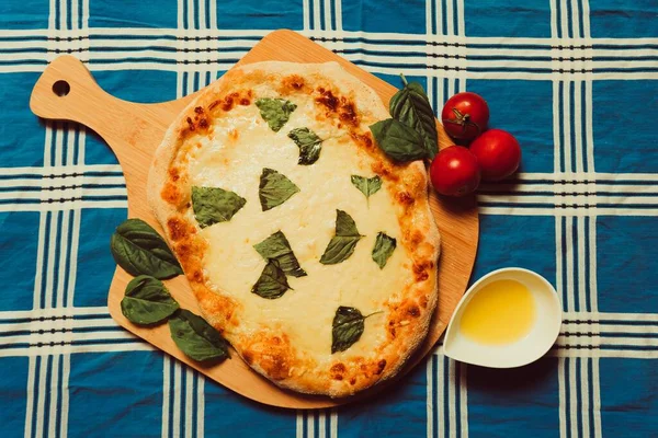 An overhead shot of a homemade pizza on a patterned surface, with marinara sauce, olive oil, fresh mozzarella, and basil leaves