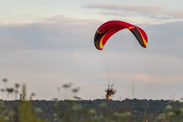 A tandem of people under the red dome of a motor paraglider takes off in the air over a green field against a blue sky. Red dome of a motor paraglider