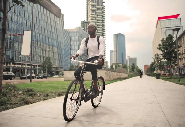 一个戴着眼镜和背包的非洲男人骑着自行车走在被现代建筑环绕的人行道上 — 图库照片