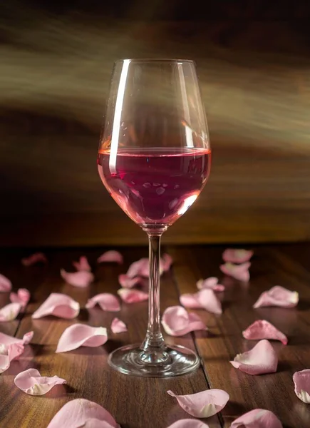 A glass of rose wine and pink rose petals around on the wooden surface