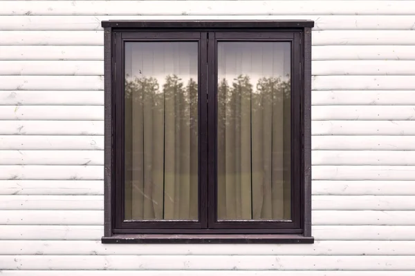 A brown wooden window in the middle of a white wooden wall with the reflection of the trees in it