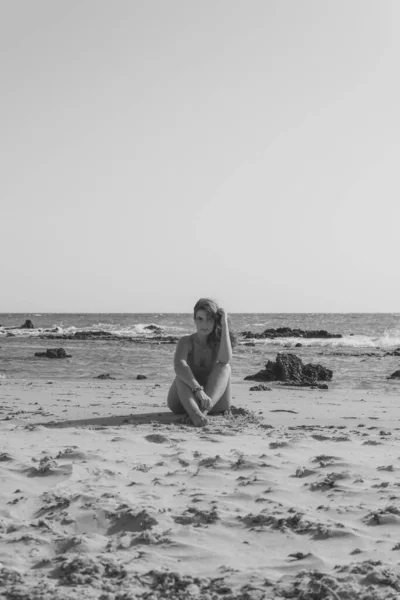 一个身穿泳衣的欧洲美女坐在沙滩上的垂直灰度照片 — 图库照片