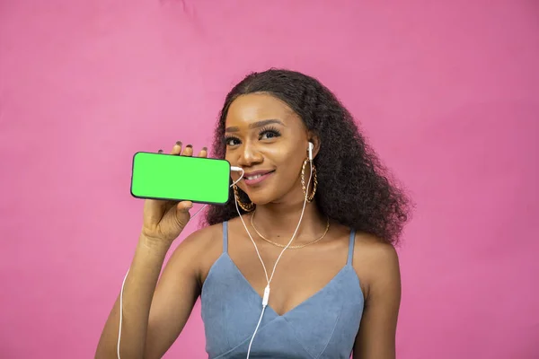 一个戴着耳机 在粉色背景上显示手机屏幕的年轻非洲妇女 — 图库照片