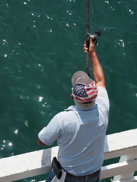 A vertical shot of a man wearing a hat with the American flag on it, fishing off a pier during daylight