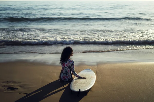 太陽の光の下でビーチに座ってサーフボードと若い女性の浅い焦点 — ストック写真