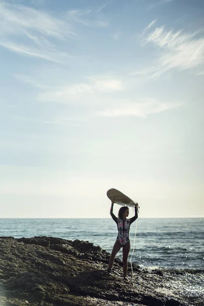 ビーチでサーフボードを持っている水着の若い女性の垂直ショット — ストック写真