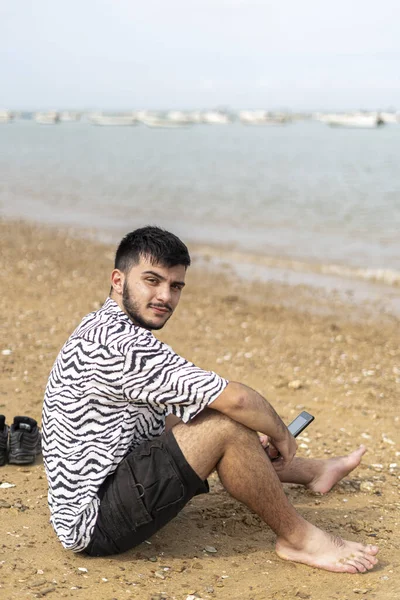 Szczęśliwy Młody Mężczyzna Smartfonem Siedzącym Plaży Pozującym Przed Kamerą — Zdjęcie stockowe