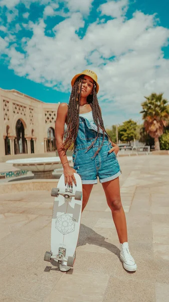 A female with dreadlocks in a bucket hat and with skateboard posing at camera
