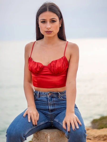 美しいです若いです女性身に着けていますセクシーな赤い作物の上とジーンズと背景に海と岩の上に座って — ストック写真
