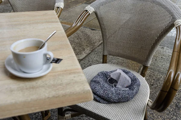 帽子とフェイスマスクを備えた椅子の横にあるカフェテーブルの上のコーヒーカップ — ストック写真