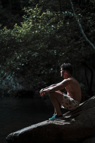 一个年轻的白人男子坐在河边的岩石上 周围环绕着美丽的绿叶 — 图库照片