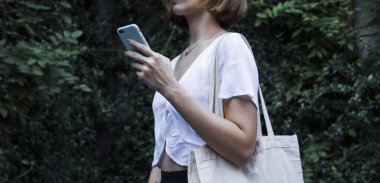 Boş beyaz alışveriş tuvali kumaş çanta ve akıllı telefon elinde model tutan ve izole edilmiş doğal arka plan şablonu olan kadının üzerinde.