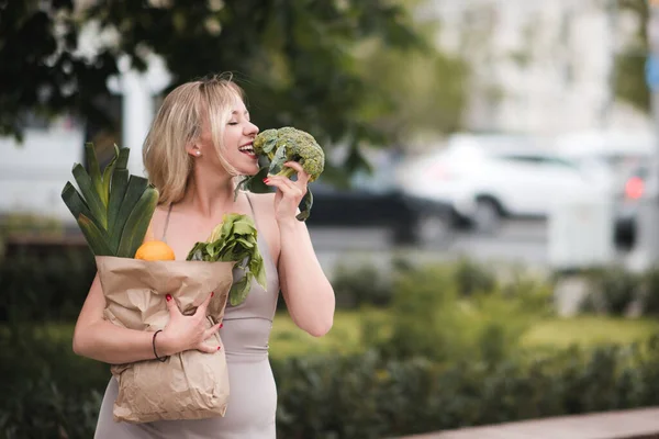 Smiling young woman 25-26 year old eating fresh green broccoli holding paper bag with food outdoors closeup. Healthy lifestyle. 20s.