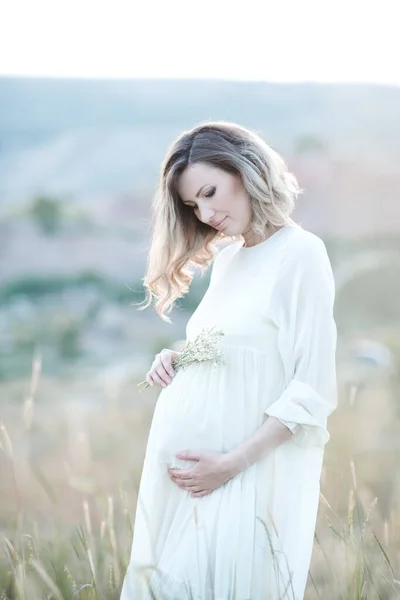 日落时分 美丽的孕妇穿着白色的连衣裙 站在户外的草地上 沐浴在阳光下 — 图库照片