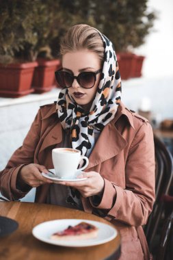 24-25 yaşlarındaki güzel bir kadın kafede kahve içiyor şık bir ceket, güneş gözlüğü ve açık havada ipek fular takıyor.. 