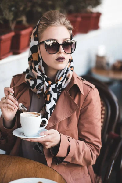Beautiful woman 24-25 year old drinking coffee in cafe wearing stylish jacket, sun glasses and silk scarf outdoors.