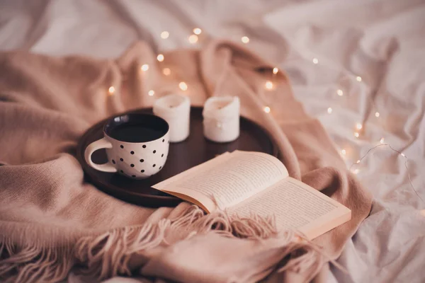Bir fincan açık kitaplı kahve ve ahşap tepside mumlar. Yatakta da örülmüş kumaş var. İyi sabahlar. Kış mevsimi. 