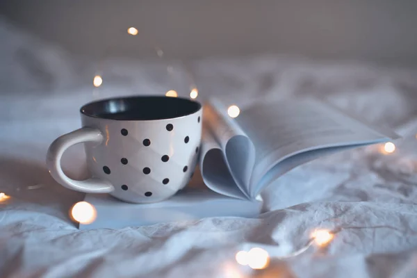 Cup of tea on open book with heart shape folded pages closeup in bed at night. Autumn season.