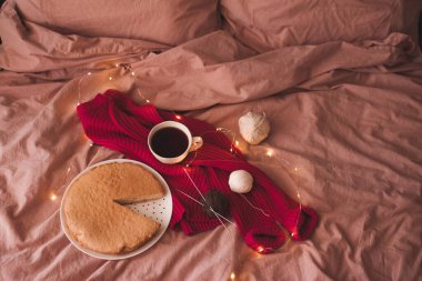 Bir fincan siyah çay, pembe örülmüş kumaş, lezzetli bir turta ve parıldayan Noel ışıklarıyla yatakta iplik. Seçici odaklanma. Kış tatili sezonu. Günaydın..