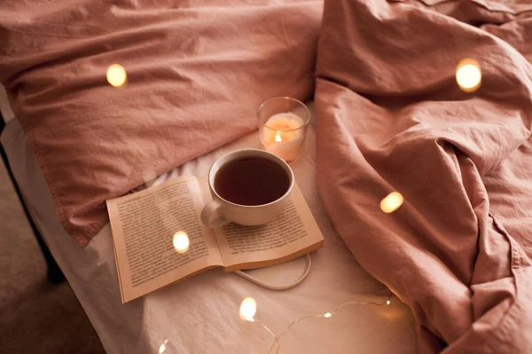 新鮮なホットコーヒーのカップは ベッドの近くに燃えるろうそくと輝くクリスマスライトと開いて紙の本に滞在します 冬休みの季節 おはようございます 選択的焦点 — ストック写真