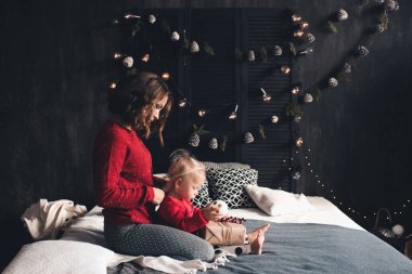 Anne, 1-2 yaşındaki bebeğini kucağında tutuyor. Kırmızı örgü süveter giyiyor. Odada Noel süsleri var. Kış tatili. Annelik. Xmas.