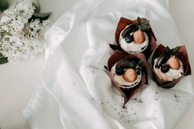 Meyveli kek kremalı beyaz bir çiçekle süslenmiştir. Üstü çikolata parçalı kurabiye ve yaban mersinli. Kompozisyon yeşil yapraklarla bitiyor. Kağıt bardağa doldurulmuş..