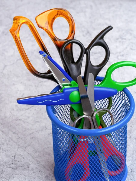 Scissors of different sizes and for different jobs are in a mesh container. Children's scissors with a wavy edge lie across the container.