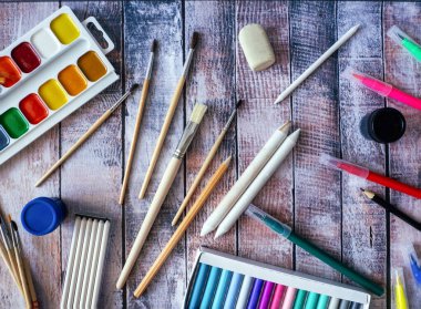 Watercolors, art brushes, pastels, felt-tip pens, and other painting tools are on a wooden surface. Creative background with tools for artwork. clipart