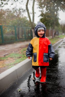 Yağmurda tek başına koşan küçük bir çocuk kalyuzhla koşar. Mavi, kırmızı ve turuncu renkli ve kırmızı çizmeli bir yağmurlukla. Mutlu çocuk. Tasasız çocukluk. Kötü hava. Yağmuru severim. sis.
